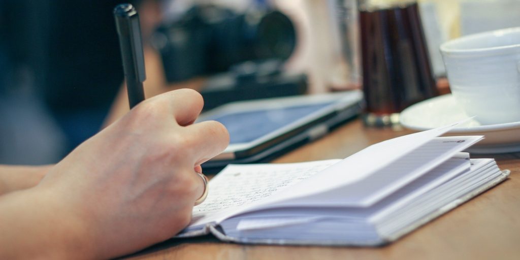 Someone holding a pen and writing on a notebook. Only their hand is visible.